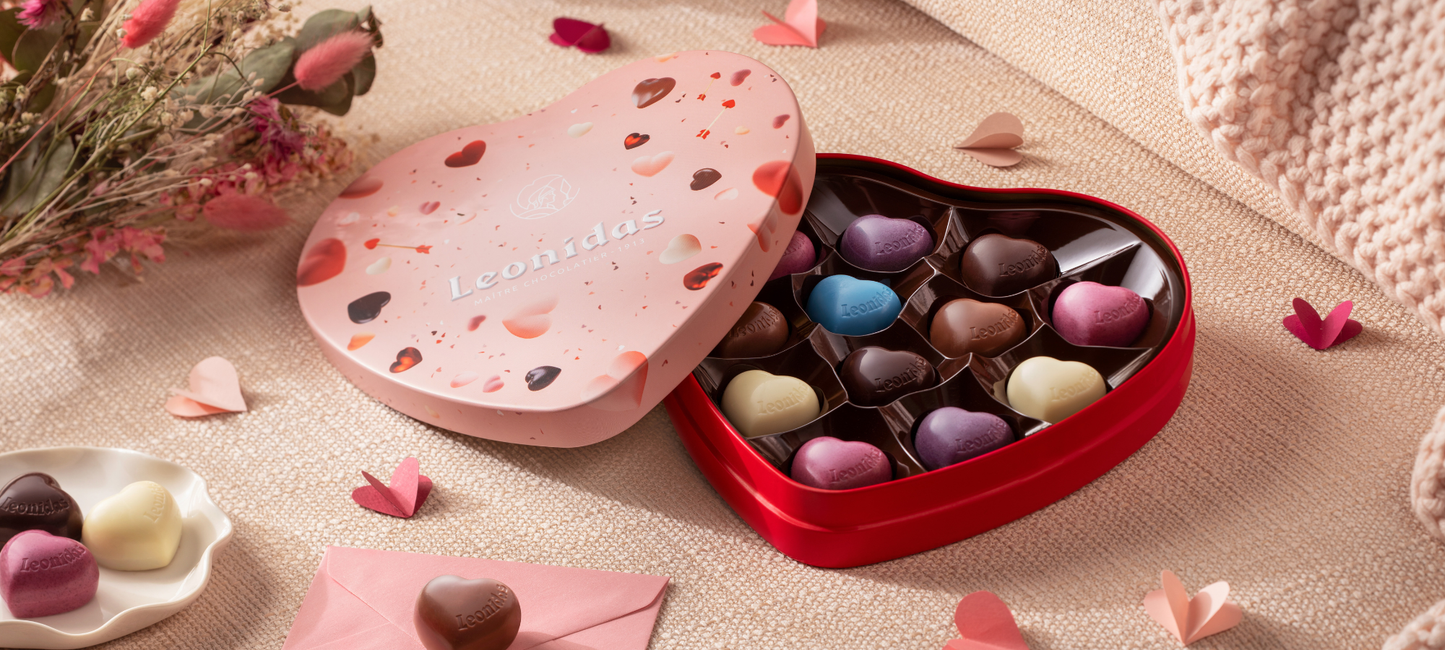 Heart-shaped chocolate box with colorful chocolates on a soft surface with flowers and petals.